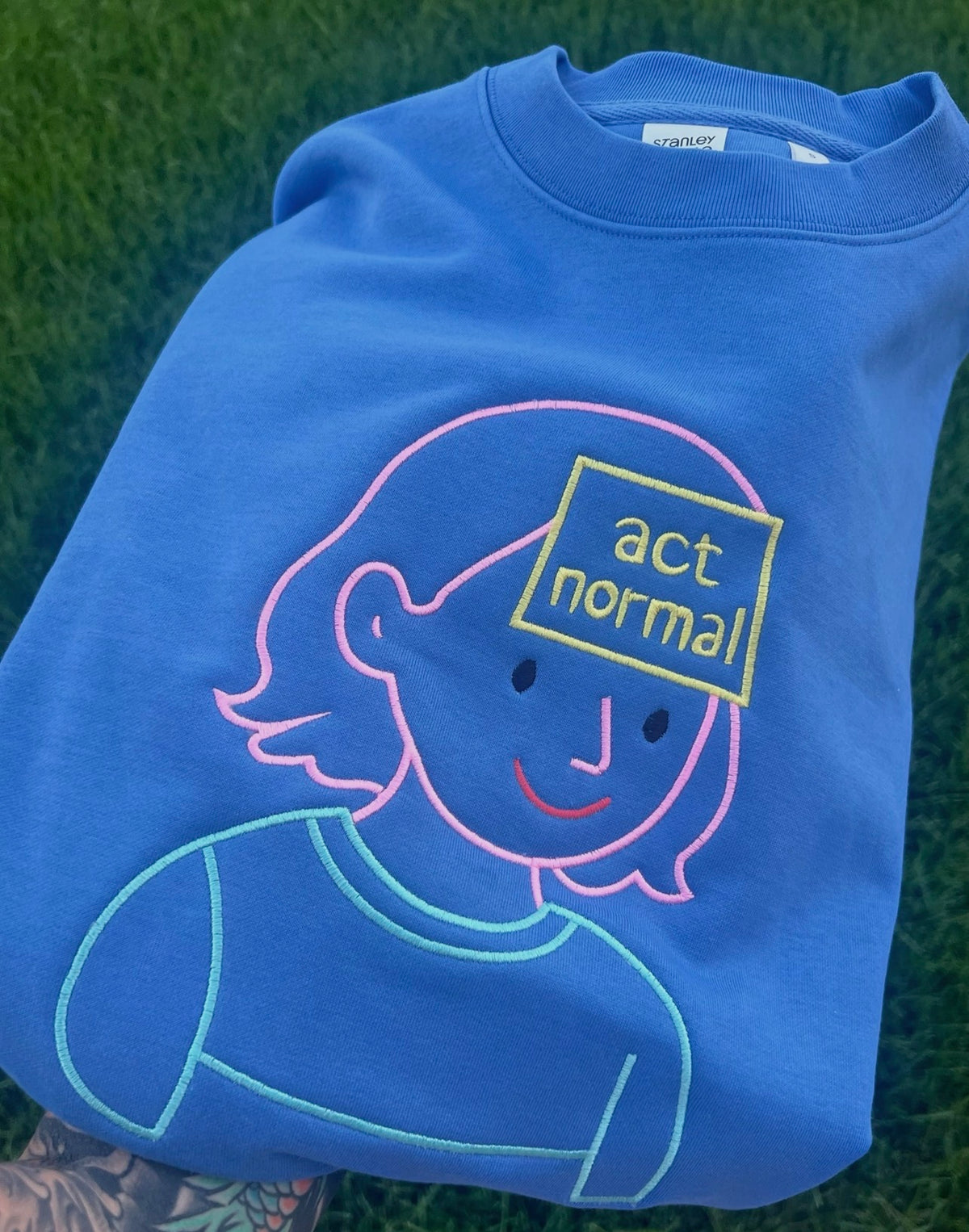Blue sweatshirt with embroidered person holding an Act Normal sign
