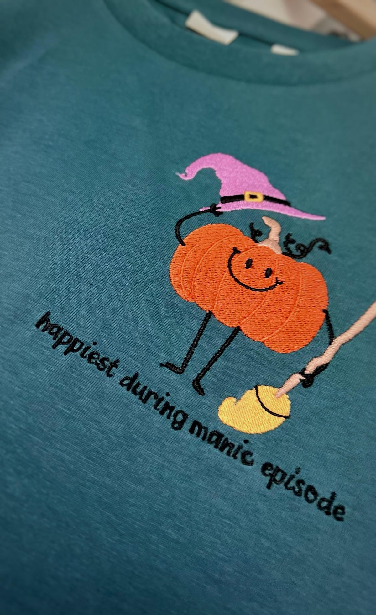Teal sweatshirt with orange pumpkin in a witch hat and Happiest During Manic Episode lettering