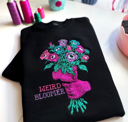 Black sweatshirt with colorful embroidered design of flowers and text 'WEIRD BLOOMER' on a white surface.